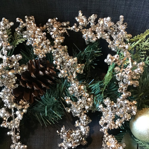 Glitter garland with shimmering champagne beads intertwined with pine greenery, pinecones, and a ball ornament against a dark gray background.  