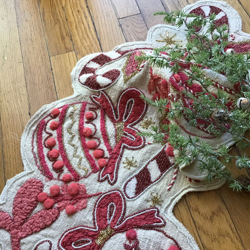 Festive cream-colored hand-embroidered table runner featuring red and pink ornaments, candy canes, and bows with red and gold beaded details and pink and red pom-poms.  Displayed on a wooden surface with greenery. 
