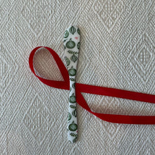 Load image into Gallery viewer, Holiday Spreaders | Four Styles, lifestyle photo of a white enameled stainless steel spreader with green ornaments and foliage design, displayed with a red ribbon on a beige textured fabric surface. 