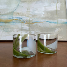 Load image into Gallery viewer, Splash Cups hand-blown clear glass set of two with abstract pistachio green and white swirled design on wooden surface against abstract background. 