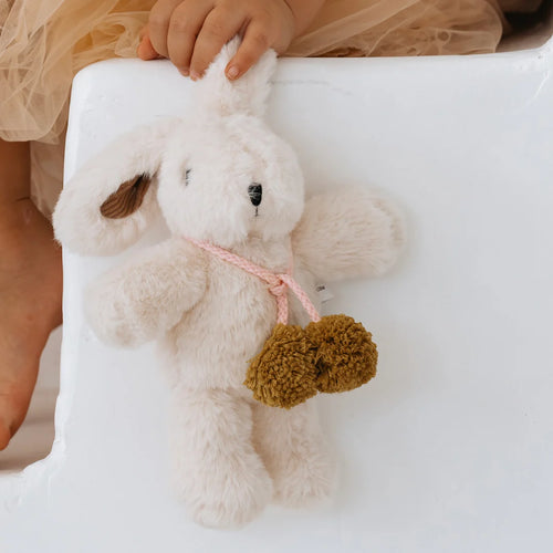 Vanilla cream-colored plush bunny toy with soft plush fabric, a pink tie with two large Australian merino wool chartreuse pom-poms, shown being held by a child wearing a beige tulle skirt,  against a white surface.  