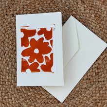 Load image into Gallery viewer, Colby Cards, lifestyle image of a white Lokta paper greeting card featuring bold abstract red-orange flower silhouettes within a white border, displayed with a white envelope on a brown woven mat.