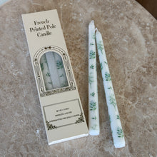 Load image into Gallery viewer, Two white taper candles with blue-and-green botanical designs on a brown marble surface, next to their packaging. 