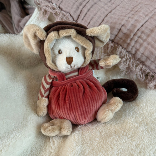 White plush bear dressed in a monkey-inspired outfit with a brown hood featuring cream-colored rounded ears, hands, and feet, a salmon-colored corduroy romper, and salmon and white striped long sleeves. Plushie is sitting on a soft white blanket with a pink textured textile in the background. A brown plush tail curls to the bears side. 