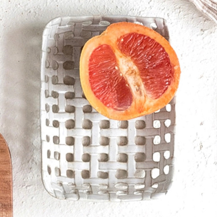 A white stoneware dish with latticework pattern, displaying a sliced citrus fruit in its basket, placed on a white textured  surface.