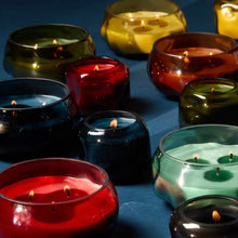 Load image into Gallery viewer, Collection of colorful lit candles in glass holders on a blue surface. Colors include green, red, blue, yellow, brown, and mint green.