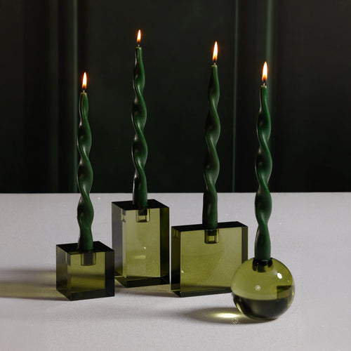 Four green twist taper candles, each lit and displayed in green crystal glass holders including a large and small square holder, a rectangular shaped holder, and a round holder, set on a white surface against a dark background. 