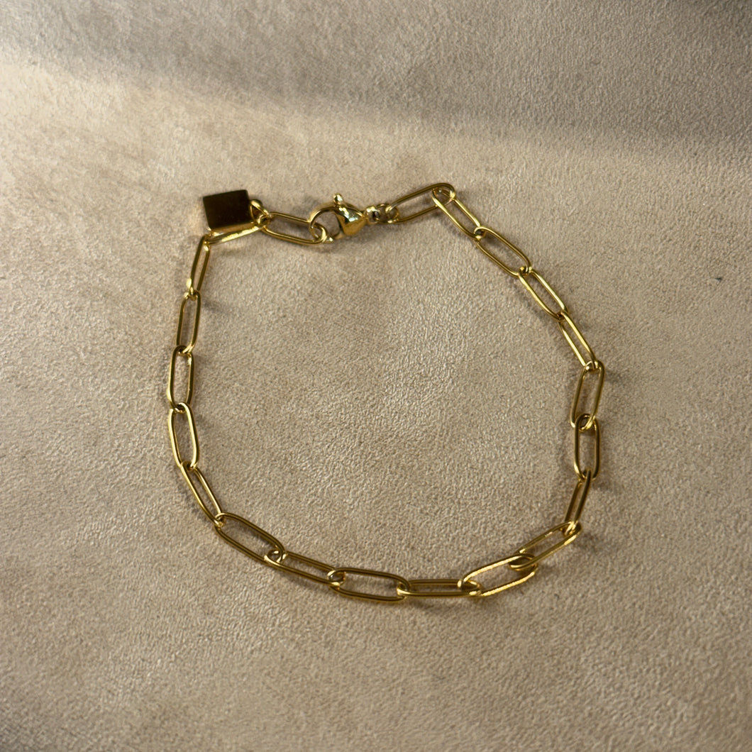 Gold-plated stainless-steel charm bracelet with elongated oval links, displayed on a soft beige surface. 