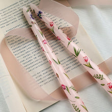 Load image into Gallery viewer, Set of two pale pink floral taper candles with tulip and wildflower designs, displayed on an open book with a sheer pink ribbon.