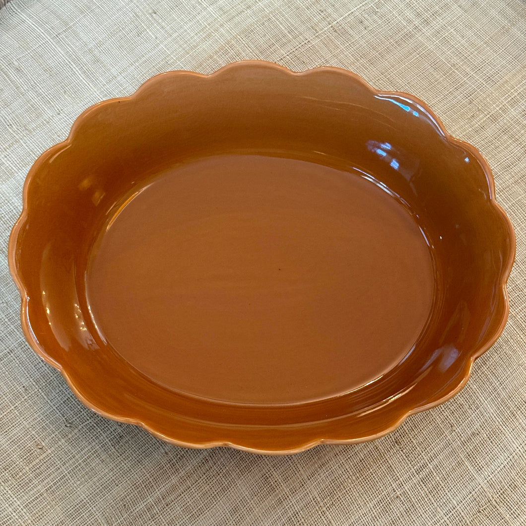 Oval terracotta-colored stoneware dish with scalloped-edges displayed on a cream-colored textured surface. 