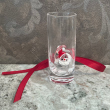 Load image into Gallery viewer, ZODAX Holiday Shot Glasses, lifestyle image of a tall clear shot glass with a thick base and a raised Santa face featuring a white beard, red hat, and red nose, resting on a gray marble surface with a red ribbon and a patterned gray background.