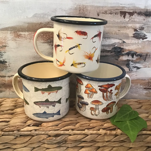 Three aluminum coffee mugs stacked on a braided basket in front of an abstract painting background. One mug has a black rim and a print of multi-colored fishing lures on it. Another has stripes of multi-colored fish and a black rim. And the other has multi-colored mushrooms and blue rim. 