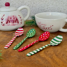 Load image into Gallery viewer, A red and white striped stainless steel spoon. A green stainless steel spoon with white polka dots. A red stainless steel spoon with white polka dots. A green and white striped stainless steel spoon. A white ceramic teapot with a red round top. Teapot has a red house, trees, snowmen, and botanical motif design on its front. A white ceramic mug with &quot;Home for the HOLIDAYS&quot; in red text. On a brown wooden surface with a green fir tree clipping. 