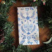 Load image into Gallery viewer, IHR lunch-sized paper napkin set with a Hanukkah design featuring blue menorah&#39;s, Stars of David, dreidels, and leafy motifs on a white background. Displayed on a wooden surface surrounded by green garland.  