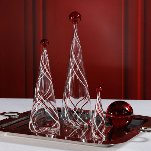 Three clear glass Christmas trees in varying heights with red and white swirling stripes, each topped with a red glass sphere. Displayed on a reflective silver tray beside a matching red glass orb, against a red background and a white surface. 
