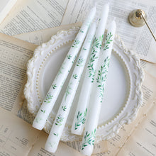 Load image into Gallery viewer, Two sets of white taper candles with assorted botanical designs, displayed on an ornate white plate over scattered paper pages.