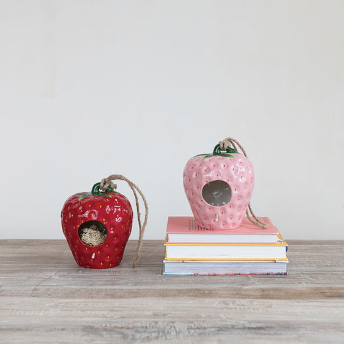 Two birdhouses that resemble strawberries, one red and one pink. The pink birdhouse is displayed on a stack of books, on a wooden surface and a white background.