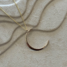 Load image into Gallery viewer, 14K gold-filled necklace featuring a large hammered crescent-moon pendant on a delicate gold chain, displayed on a textured tan surface. 