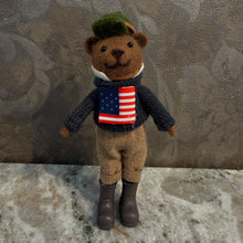 Load image into Gallery viewer, Preppy Bears Ornaments, lifestyle image of a brown teddy bear wearing a dark green cap, navy knit sweater with an American flag design, white collar, beige trousers, and dark boots, displayed on a gray stone surface against a gray patterned background.