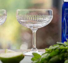 Load image into Gallery viewer, Coupe glass with line detailing. In the foreground is a bundle of mint and two lime wedges. The background is blurred nature. A blue bottle is to the side.