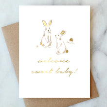 Load image into Gallery viewer, Greeting card with gold bunnies and "welcome sweet baby!" script text, with a brown kraft envelope on a marble surface.