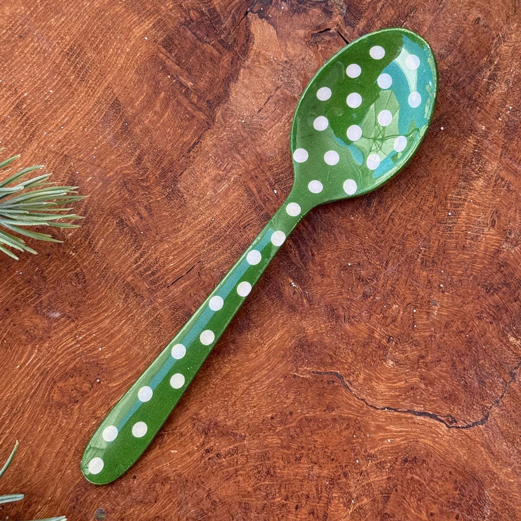 A green stainless steel spoon with white polka dots displayed on a wooden surface  with a small sprig of evergreen in the corner. 