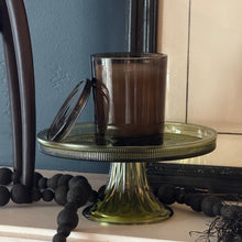 Load image into Gallery viewer, Smoky glass candle holder with candle on an olive -colored glass cake stand. Candleholder lid is propped against the holder.
 Cake stand has a ribbed lip that is partially black. The stand is on a white surface garnished with a strand of black bead garland and a tall black object. Against a dark blue background with a white picture in a black frame.