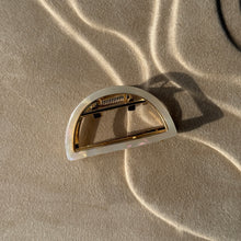 Load image into Gallery viewer, Ivory half-moon metal claw clip with gold hardware displayed on a beige textured surface with soft curved light reflections.