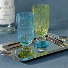 Load image into Gallery viewer, ZODAK Antibes Mimosa Bellini glasses in translucent blue bubble glass with a green pedestal base and translucent yellow-green bubble glass with a blue pedestal base, displayed on a mirrored tray beside a tequila bottle against a blue background.