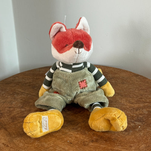 Plush fox with pumpkin orange and white fur, black stitched eyes, a brown nose, yellow hands, and yellow floppy feet. The fox is wearing a green-and-white striped shirt, and moss-colored overalls with a coral patch detail. Fox is sitting upright on a wooden surface, and against a white background. 