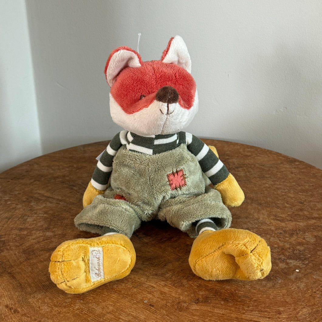 Plush fox with pumpkin orange and white fur, black stitched eyes, a brown nose, yellow hands, and yellow floppy feet. The fox is wearing a green-and-white striped shirt, and moss-colored overalls with a coral patch detail. Fox is sitting upright on a wooden surface, and against a white background. 