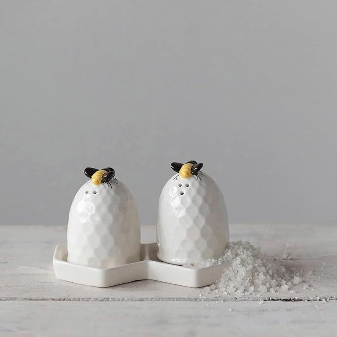 A pair of white beehive-shaped salt and pepper shakers with a black and yellow bee  on top, sitting on a white tray. Shakers and tray on a white wooden surface with a pillar of salt . Against a white background.