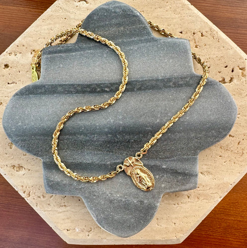 Gold rope chain necklace with a stamped Mother Mary pendant, displayed on a quatrefoil-shaped gray stone tile layered over a beige travertine slab on a wooden surface.  