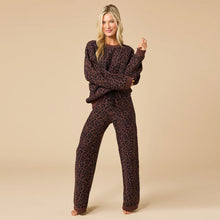 Load image into Gallery viewer, Person standing and smiling in an espresso-colored lounge set featuring a crewneck sweater and matching wide-leg pants with a black leopard print pattern. The relaxed-fit outfit includes ribbed cuffs and hems, shown against a soft beige background. 