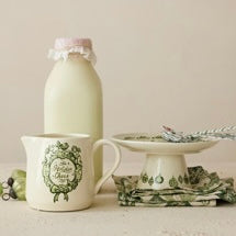 Load image into Gallery viewer, Holiday Pedestal, lifestyle image of a cream stoneware pedestal with green holiday illustrations and a flared base, styled with silver-toned utensils beside a decorated mug, a pale bottle, and green folded fabric napkins on a light tabletop.