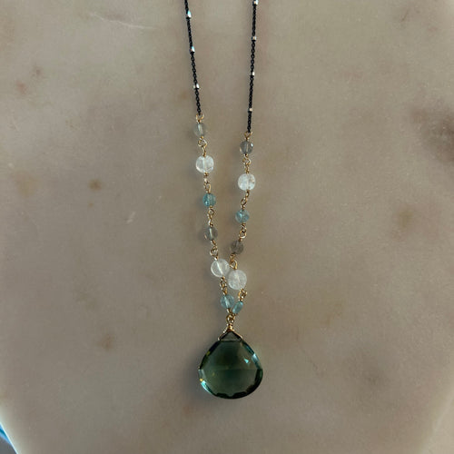 Black chain necklace that transitions to linked grey, white, and blue beads down to a blue-green teardrop gem in the center. White marble background. 