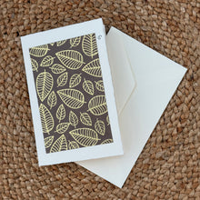 Load image into Gallery viewer, Colby Cards, lifestyle image of a white Lokta paper greeting card featuring pale gold leaf outlines on a dark gray rectangular panel, displayed with a white envelope on a brown woven mat.