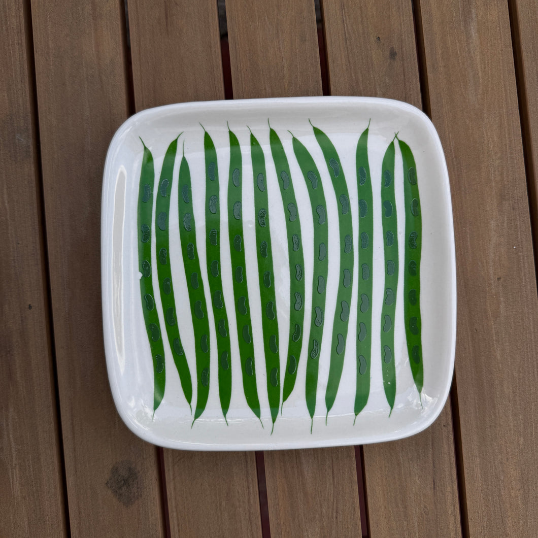 Square plate with hand painted green beans on a wooden slat surface,