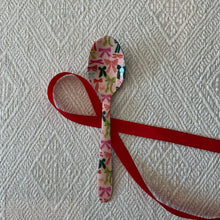 Load image into Gallery viewer, Enameled Holiday Spoons | Four Styles, lifestyle photo of a stainless steel spoon with a pale pink enamel background and a pattern of multicolored bows, displayed with a red ribbon on a cream textured fabric surface. 