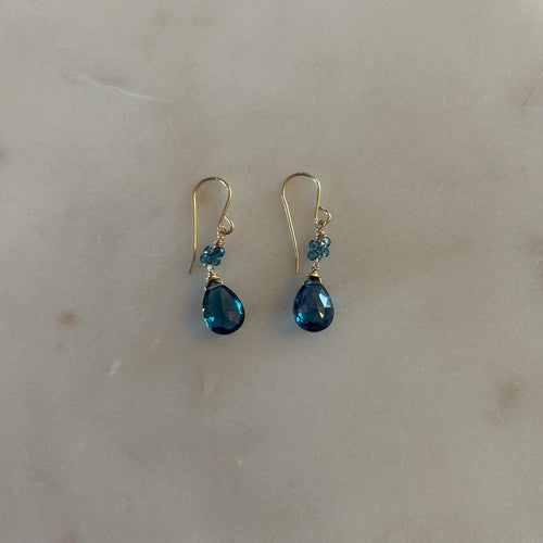 A pair of gold drop earrings with four dark blue beads and a blue teardrop gem. White marble background. 