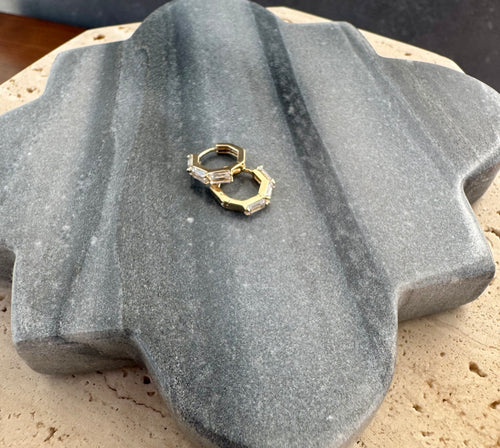 Pair of gold octagonal huggie earrings accented with clear baguette-cut crystals, displayed on a quatrefoil-shaped gray stone tile layered over a beige travertine slab. 