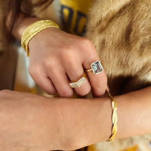 Load image into Gallery viewer, IP gold-plated stainless steel twisted snake-chain bracelet shown worn on model's wrist, with the other hand gently touching it while wearing two rings: a green tourmaline ring and a gold ring with diamond-like details. A multi-strand gold bracelet and soft tan faux fur appear in the background. 