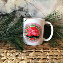 Load image into Gallery viewer, White mug with a green and pink print that reads &quot;Merry Christmas from Kensington&quot;. Background is an abstract painting and pine needles. 