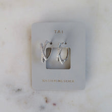 Load image into Gallery viewer, Sterling silver huggie earrings featuring an X-shape, crystal-accented design, shown on a light gray jewelry card with "TAI" in gold text and displayed on a white marble surface. 