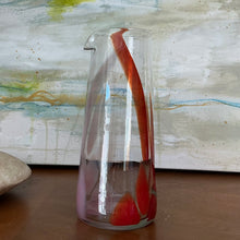 Load image into Gallery viewer, Tall clear glass pitcher with light pink and red elongated accents and pour spout, displayed on wooden surface in front of abstract painting. 