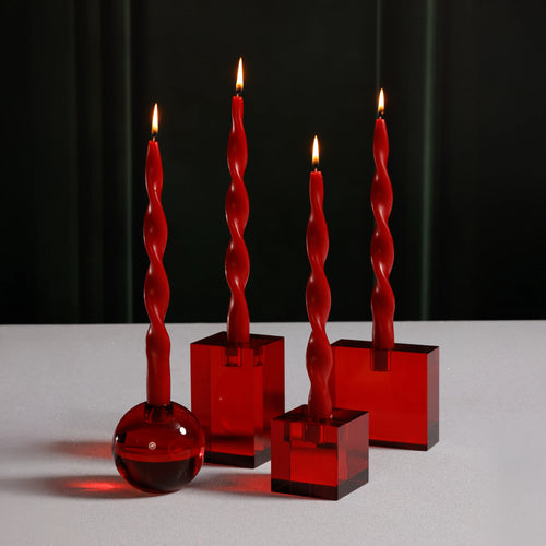 Four crimson red twist taper candles, each lit and displayed in red crystal glass holders including a large and small square holder, a rectangular holder, and one round holder, set on a white background against a dark background. 