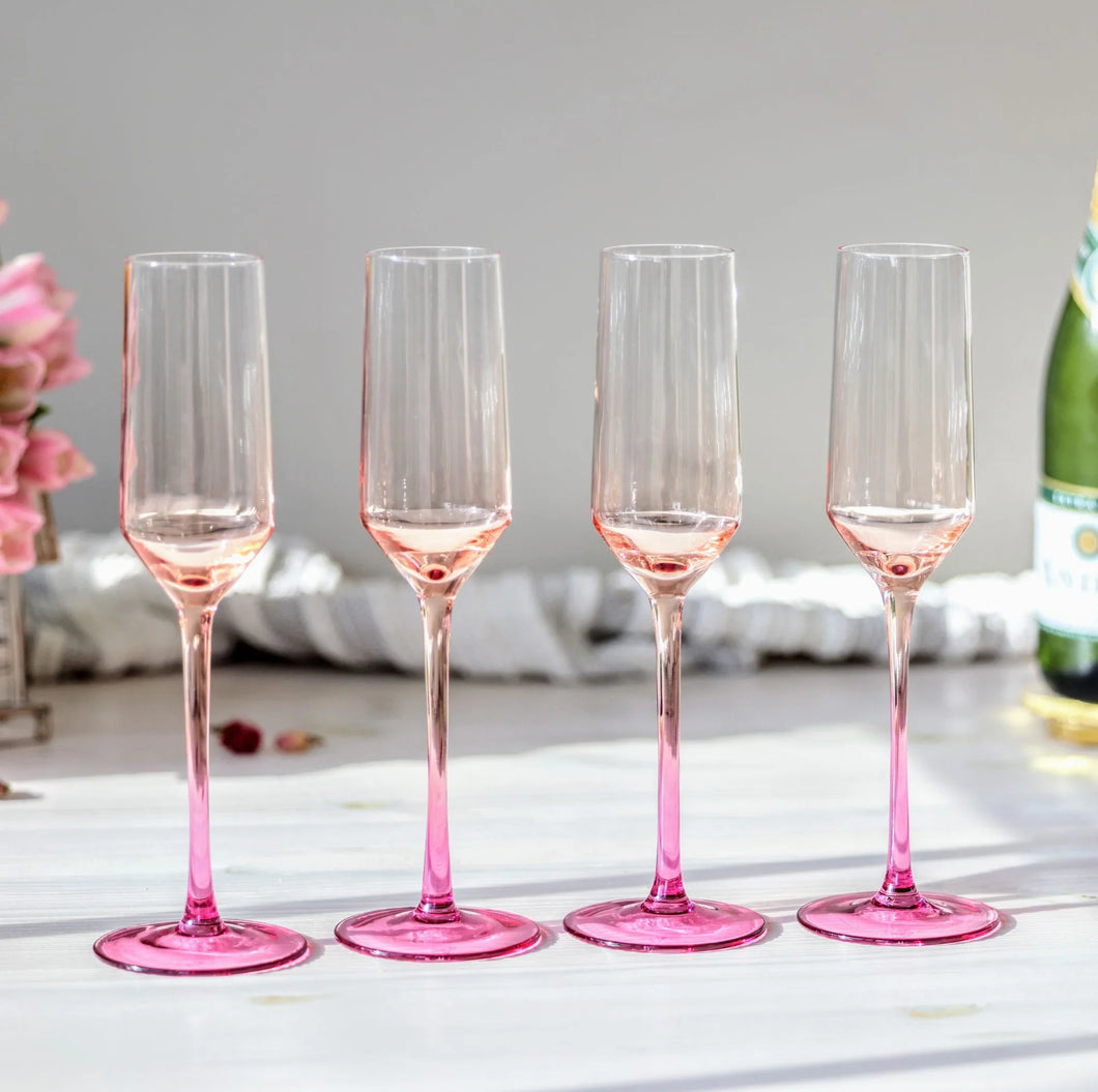Set of four hand-blown crystal champagne flutes with pink gradient stems and bases, arranged in a row on a light wooden surface with soft floral accents on the left side, displayed in front of a gray background. 