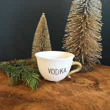 Load image into Gallery viewer, A white teacup ornament with a gold glitter handle, a clear footed bottom,  and &quot;VODKA&quot; in black text. On a wooden surface  with a green fir tree cutting and two frosted decorative Christmas trees. Against a dark background. 