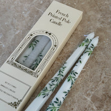 Load image into Gallery viewer, Two white taper candles with green botanical designs on a brown marble surface, next to their packaging. 