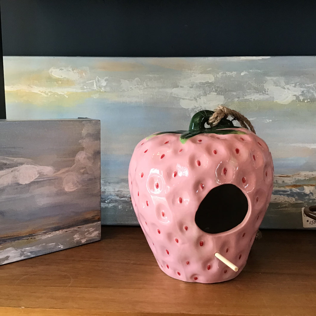 A ceramic pink birdhouse with a tied rope for hanging and a wooden perch. Two paintings of an ocean and sky in the background. Wooden surface . 